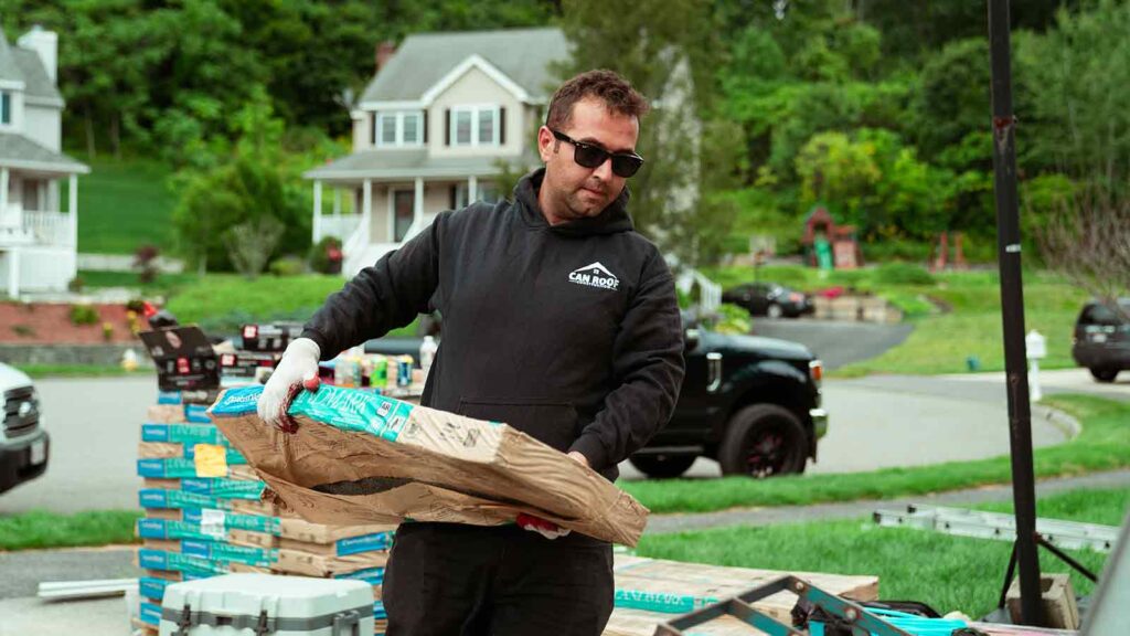 A roofing contractor wearing a black hoodie with "CAN ROOF" branding, sunglasses, and gloves