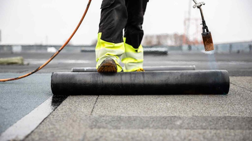Professional roofer applying tar with blowtorch on new roofing felt for waterproofing a flat roof construction site