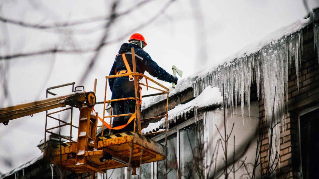 Utility worker in a safety harness and red helmet removing large icicles from a snow-covered roof using a lift in winter