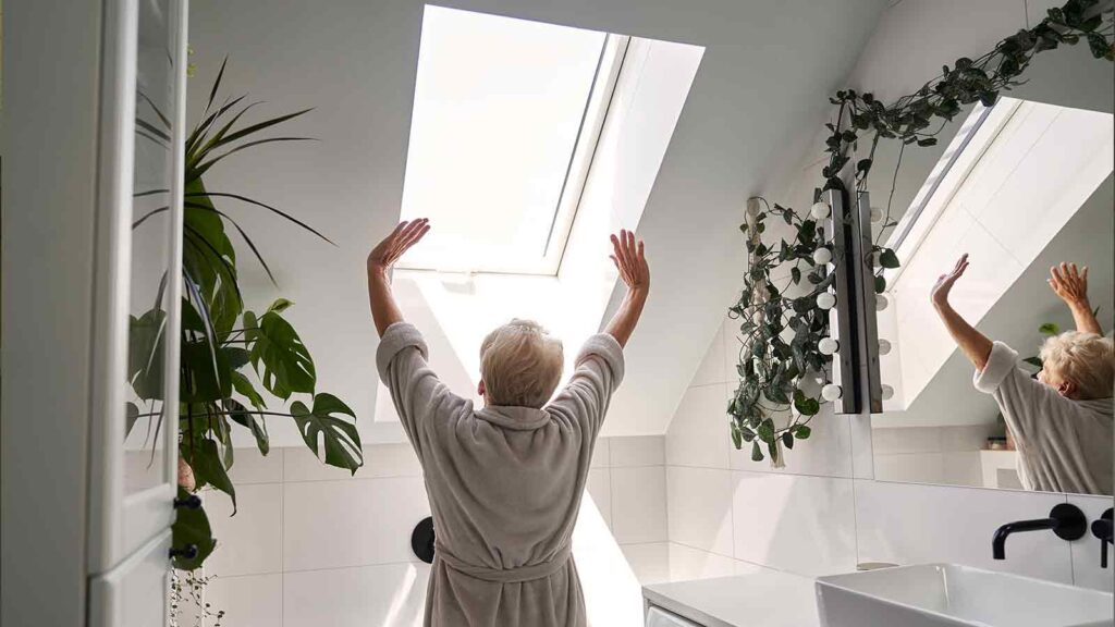 Senior woman stretching by skylight in a white bathroom with plants, washer, and vanity mirror